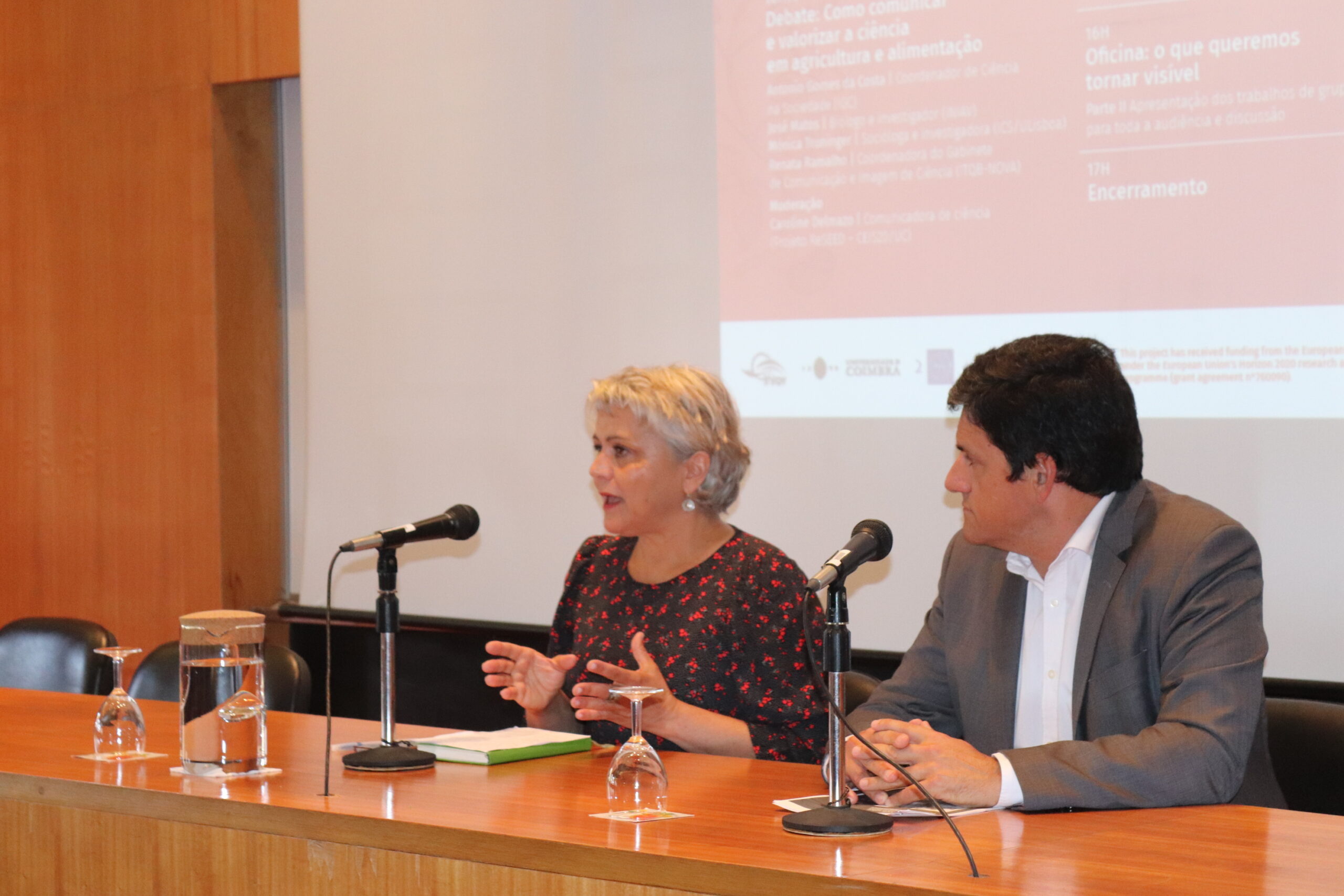 ReSEED's principal investigator Dulce Freire speaks into the microphone alongside INIAV's president, Nuno Canada, at the opening session of the workshop.They are seated at the table on the stage, the workshop poster is projected onto the background.