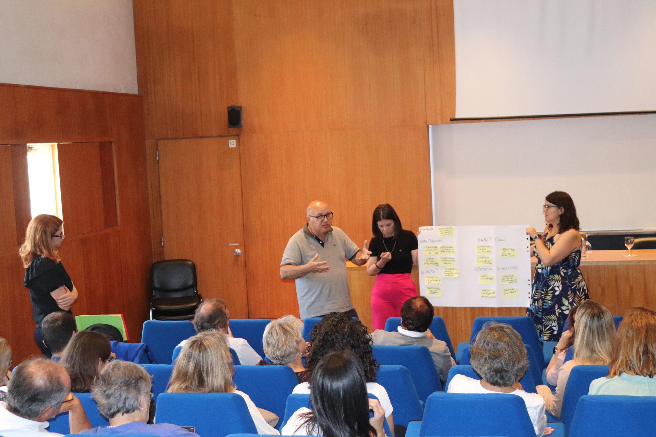 INIAV researcher José Matos presents the work of his group in the practical exercise "What do we want to make visible? He is standing in front of the audience in the auditorium. The workshop facilitators, Rita Neves and Ana Santos-Carvalho, hold an A0 sheet of paper supporting the presentation.