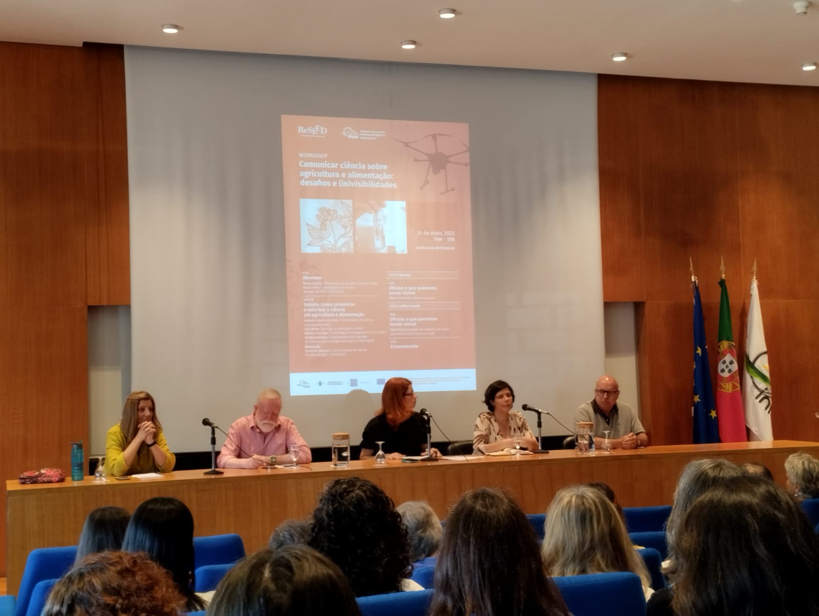 Debate table on stage: in the background, the projection of the workshop poster. From left to right, seated: Mónica Truninger, Antonio Gomes da Costa, Caroline Delmazo, Renata Ramalho and José Matos. You can also see part of the audience from the back, the auditorium chairs are blue.